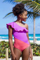 A child wearing a one-piece swimsuit with a one-shoulder design in purple and red colors, standing near a beach setting.